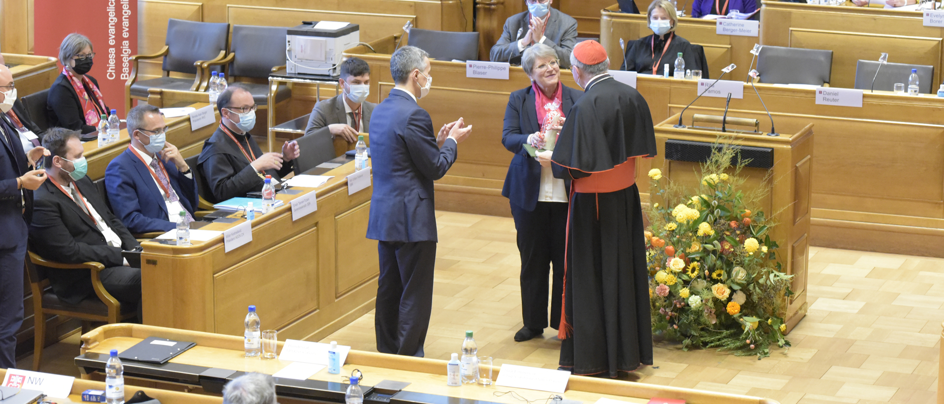 EKS-Präsidentin Rita Famos überreicht Kardinal Parolin eine Kerze und einen Heks-Spendengutschein als Geschenk, Bundesrat Cassis applaudiert.