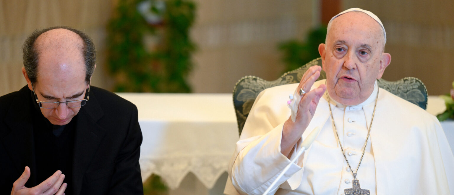Papst Franziskus spricht am 26. November 2023 im Sitzen. An seiner rechten Hand ist ein Infusionszugang zu sehen. Links von ihm Prälat Paolo Braida. | KNA